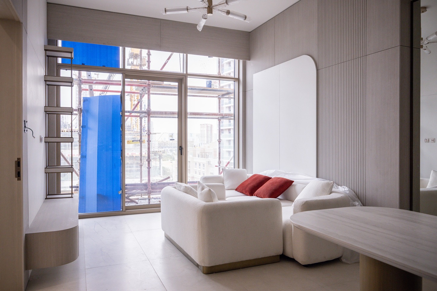 Pearl House By Imtiaz A modern living room features a white sofa with red cushions, a wooden table, and large windows revealing construction outside.